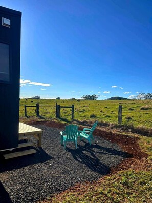 A small building with a table and chairs outside, a gravel path, and a fenced field with cows in the distance.