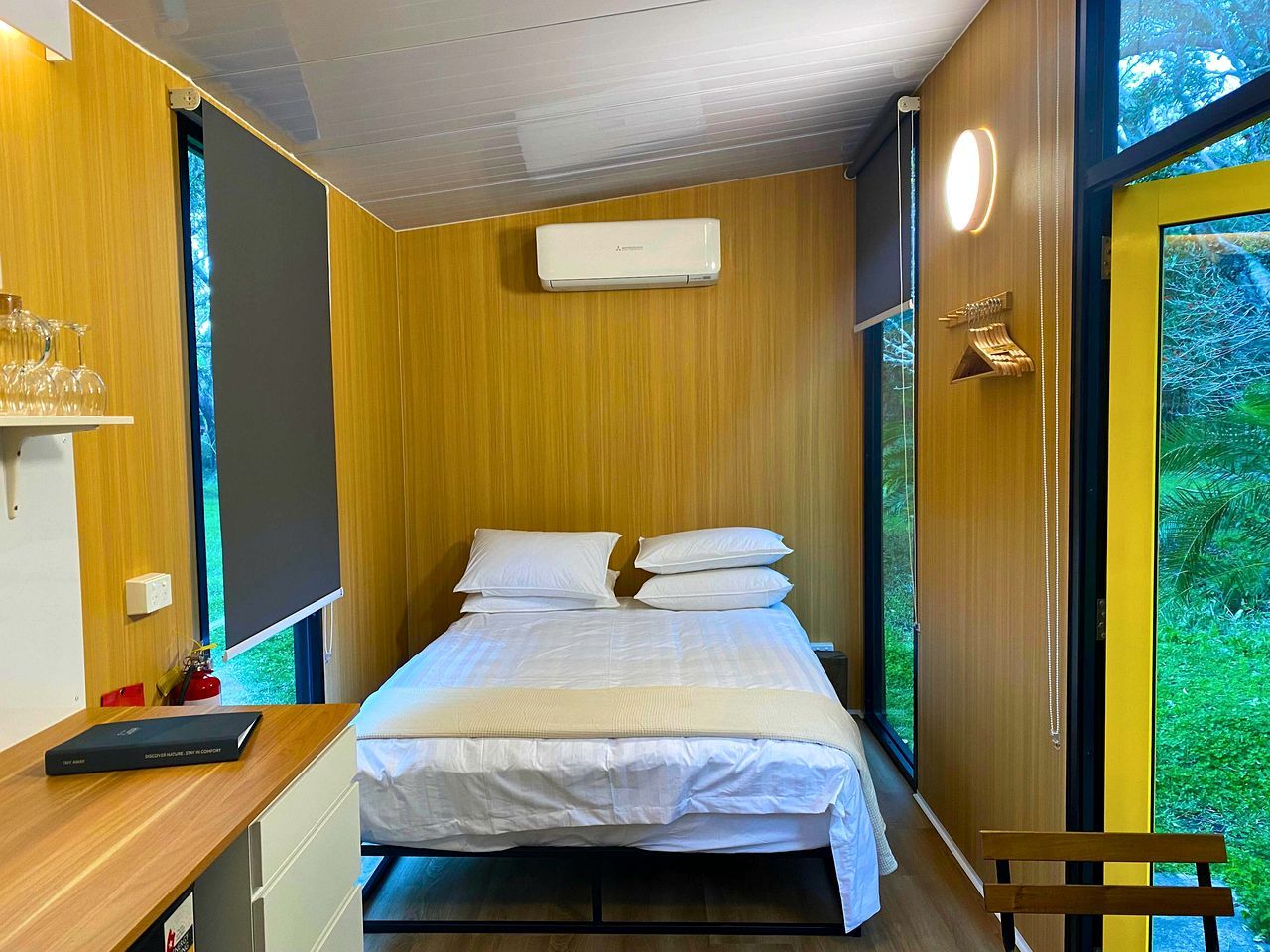 A compact room with a bed, a bedside table, a small desk, and a window with a view of greenery.