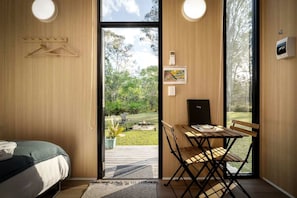 A room with a bed, a dining table with chairs, a small black cabinet, and a view of a garden through a glass door.