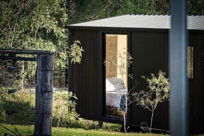 Una cabana petita de color fosc, amb un llit i una porta ben visibles, envoltada de vegetació i una tanca.