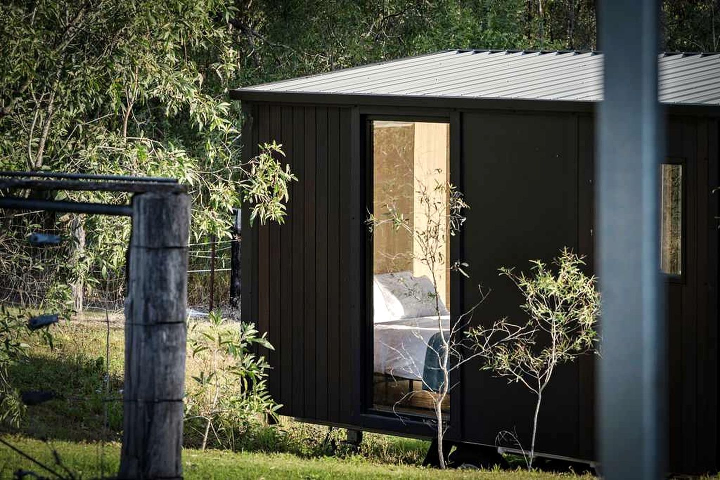 Eine kleine, dunkel gefärbte Holzhütte mit einem sichtbaren Bett und einer Tür, umgeben von viel Grün und einem Zaun.