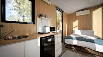 A compact bedroom with a single bed, a built-in desk with a microwave, a sink, and a window with a view of trees.