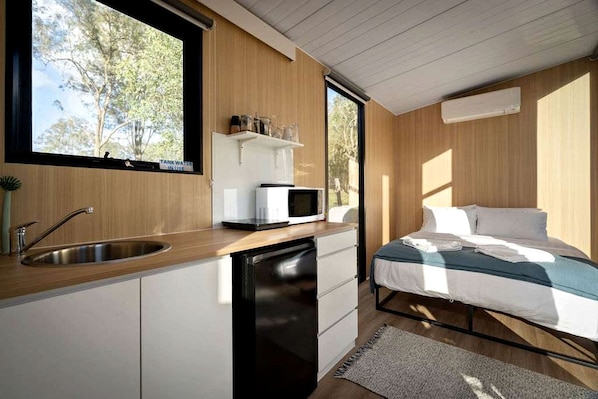 A compact bedroom with a single bed, a built-in desk with a microwave, a sink, and a window with a view of trees.