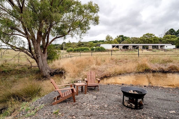 A rustic outdoor seating area with wooden chairs and a fire pit, surrounded by dry grass and a large tree.