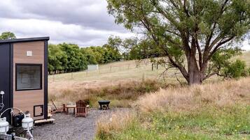 A small cabin with a gravel patio, outdoor seating, and a large tree nearby.