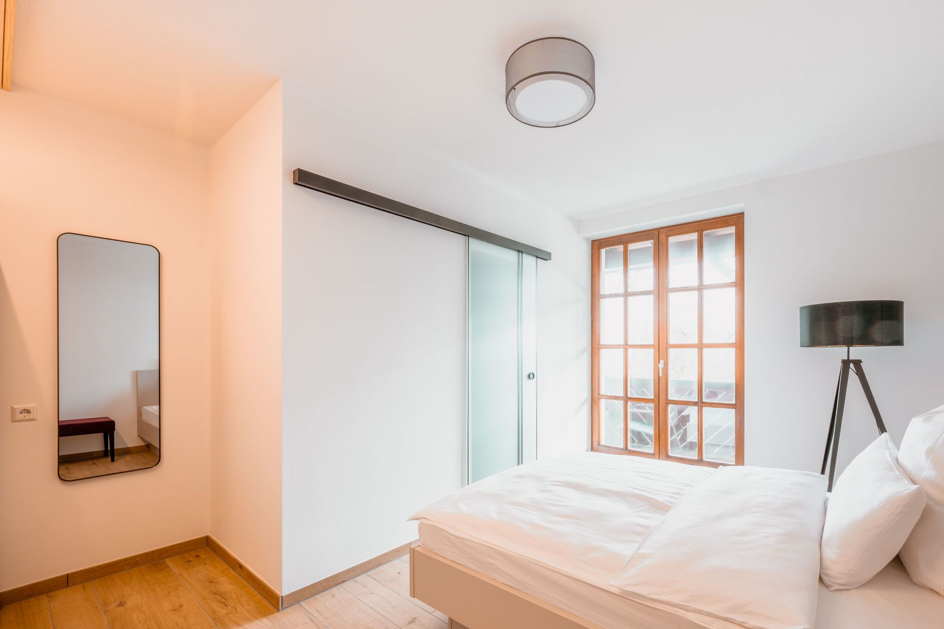 Ein Schlafzimmer mit Bett, Nachttischlampe, einem Fenster mit Blick, einer Tür und einem Spiegel an der Wand.