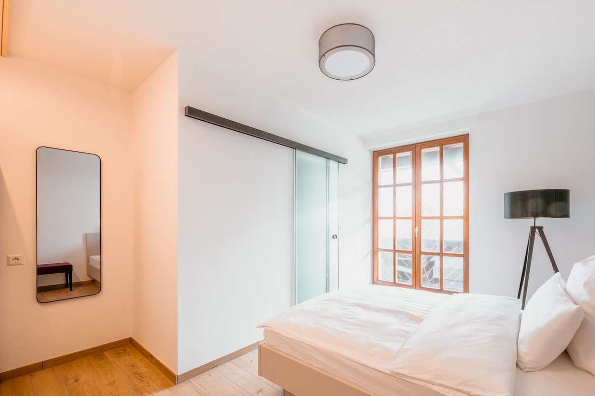 A bedroom with a bed, a bedside lamp, a window with a view, a door, and a mirror on the wall.
