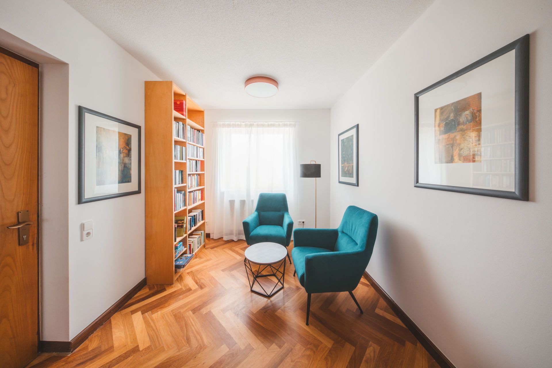 A modern living room with a wooden floor, a bookshelf, two teal armchairs, a small round coffee table, and framed artwork on the walls.