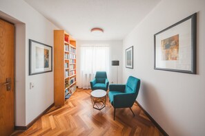 A modern living room with a wooden floor, a bookshelf, two teal armchairs, a small round coffee table, and framed artwork on the walls.