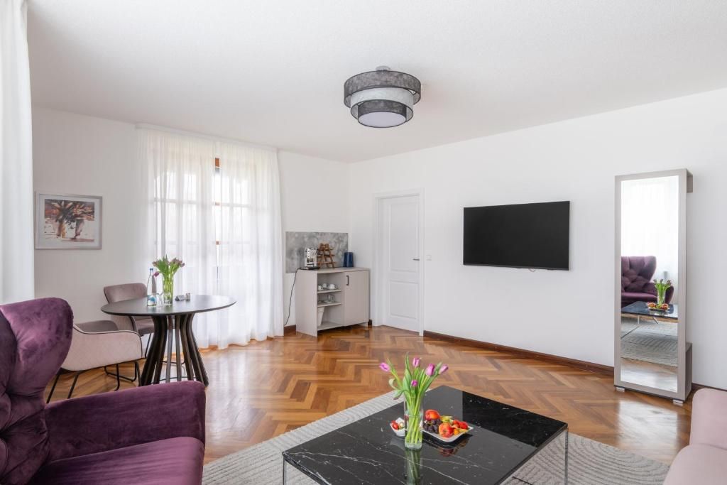 Ein modernes Wohnzimmer mit einem schwarzen Couchtisch, violetten Sesseln, einem Essbereich mit rundem Tisch, einer Küchenzeile und einem großen Spiegel.