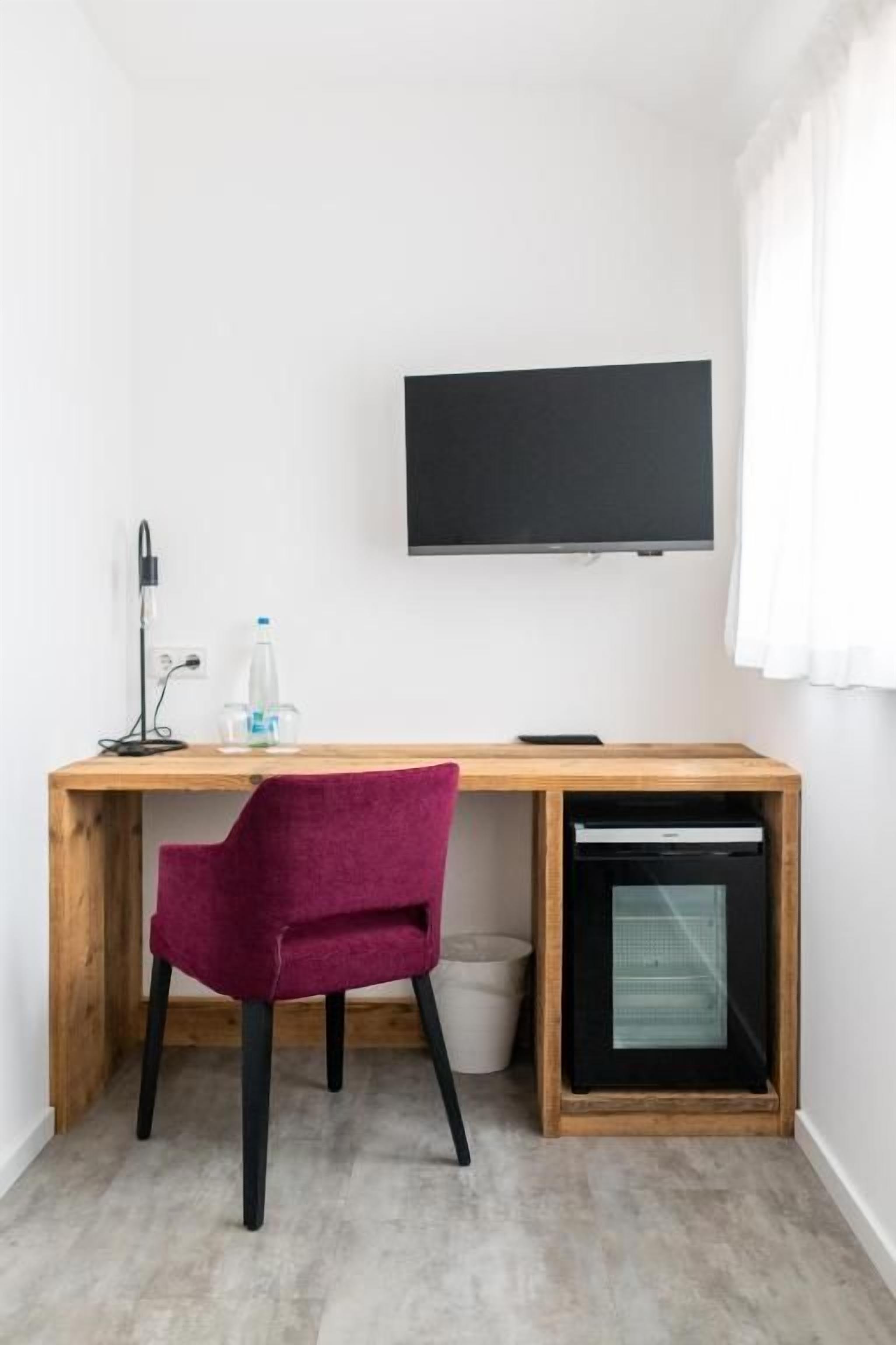 Ein Zimmer mit einem Holztisch, einem violetten Sessel, einem an der Wand montierten Fernseher und einem kleinen Fenster.
