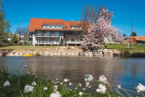 A multi-story residential building with a red roof, situated beside a calm body of water, with a flowering tree in the foreground.