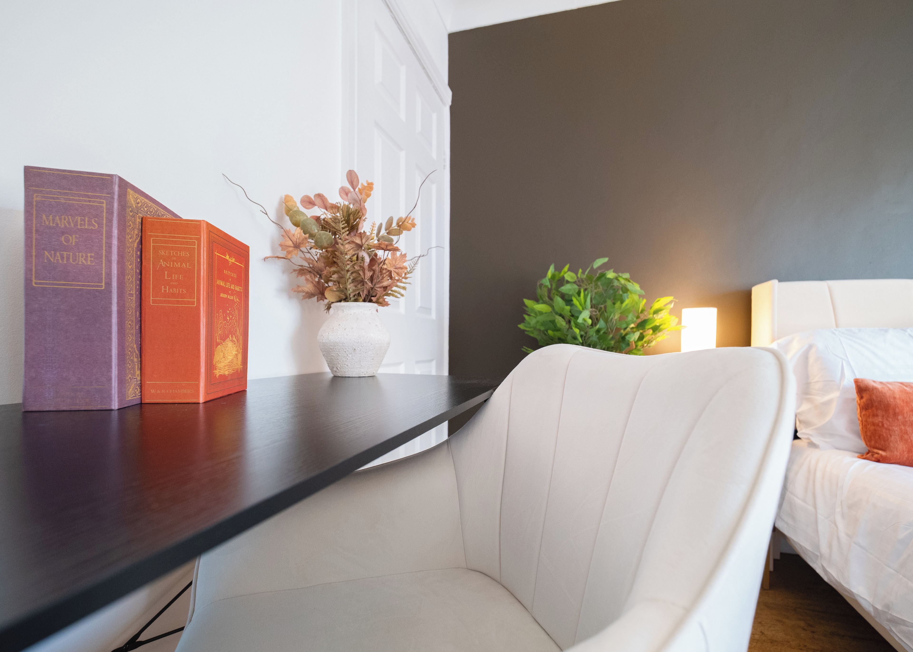 A modern room with a dark wall, a white bed, a white chair, and a nightstand with books and a vase of dried flowers.