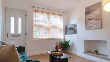 A living room with a green sofa, a coffee table with a bottle and glasses, a painting on the wall, and a window with blinds.