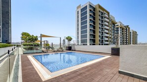 Una piscina en la azotea con cubierta de madera, rodeada de edificios modernos y algo de vegetación.
