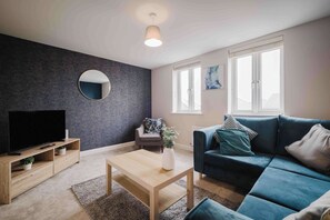 Una sala moderna con pared oscura y texturizada, un espejo redondo, un televisor de pantalla plana, una mesa de centro de madera, un sofá azul y una ventana con persianas.