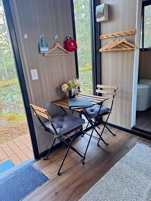 A modern room with a wooden floor, a small table with two chairs, a door with a coat rack, and a view of greenery outside.