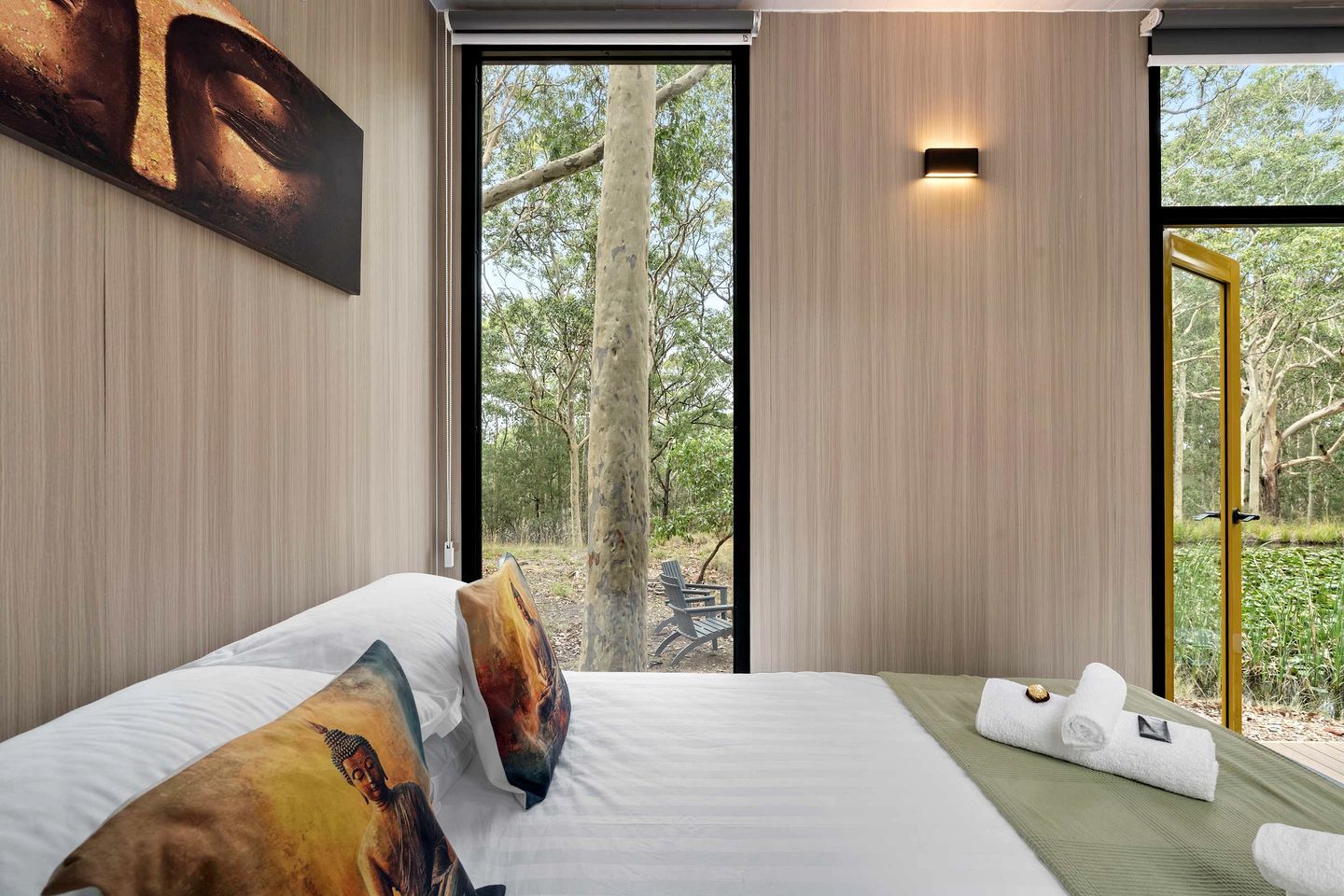 A hotel room with a bed, pillows, a towel, and a window with a view of trees.