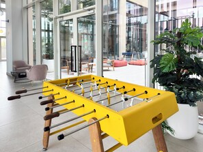 A foosball table in a modern indoor setting with glass doors leading to an outdoor seating area. - Rentico 1BR Dubai Hills Collective (Dubai)