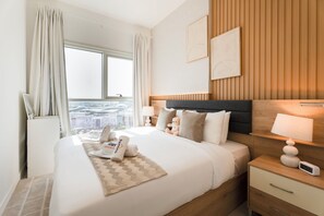 A hotel room with a large bed, a nightstand, a bedside table with a lamp, a digital clock, and a view of the city through the window. - Rentico 1BR Dubai Hills Collective (Dubai)