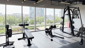 Une salle de sport équipée de divers appareils d’exercice, dotée de grandes fenêtres et offrant une vue sur des bâtiments et des espaces verts.