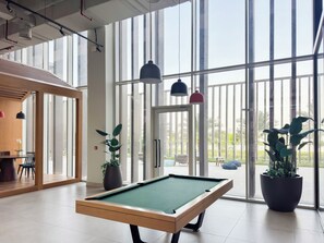 A pool table in a room with large windows, a door leading outside, and potted plants. - Rentico 1BR Dubai Hills Collective (Dubai)