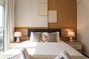 Une chambre d’hôtel avec un lit, deux tables de chevet, un ours en peluche et une fenêtre avec des rideaux.