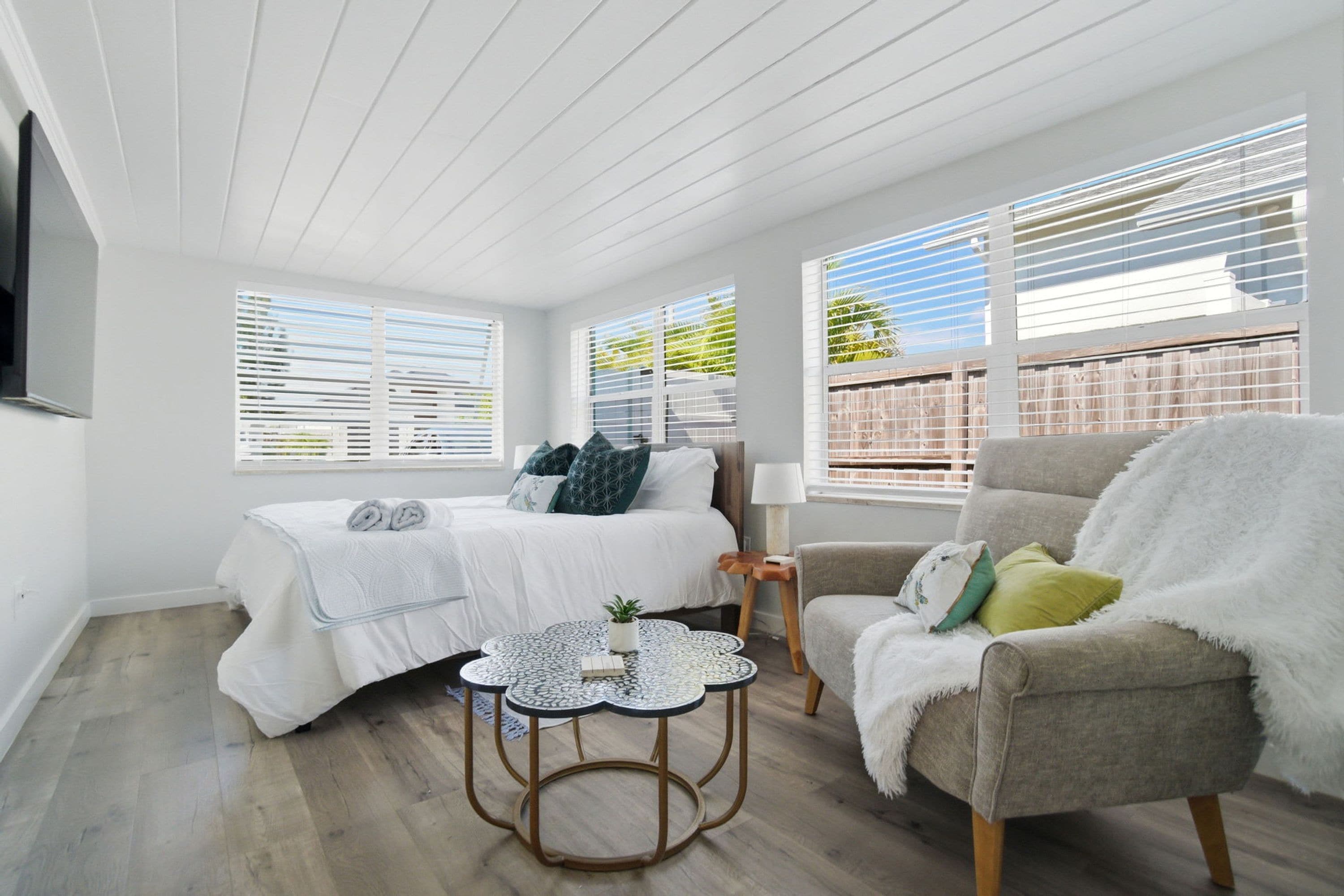 A bedroom with a bed, a chair, a small table, and large windows with blinds.