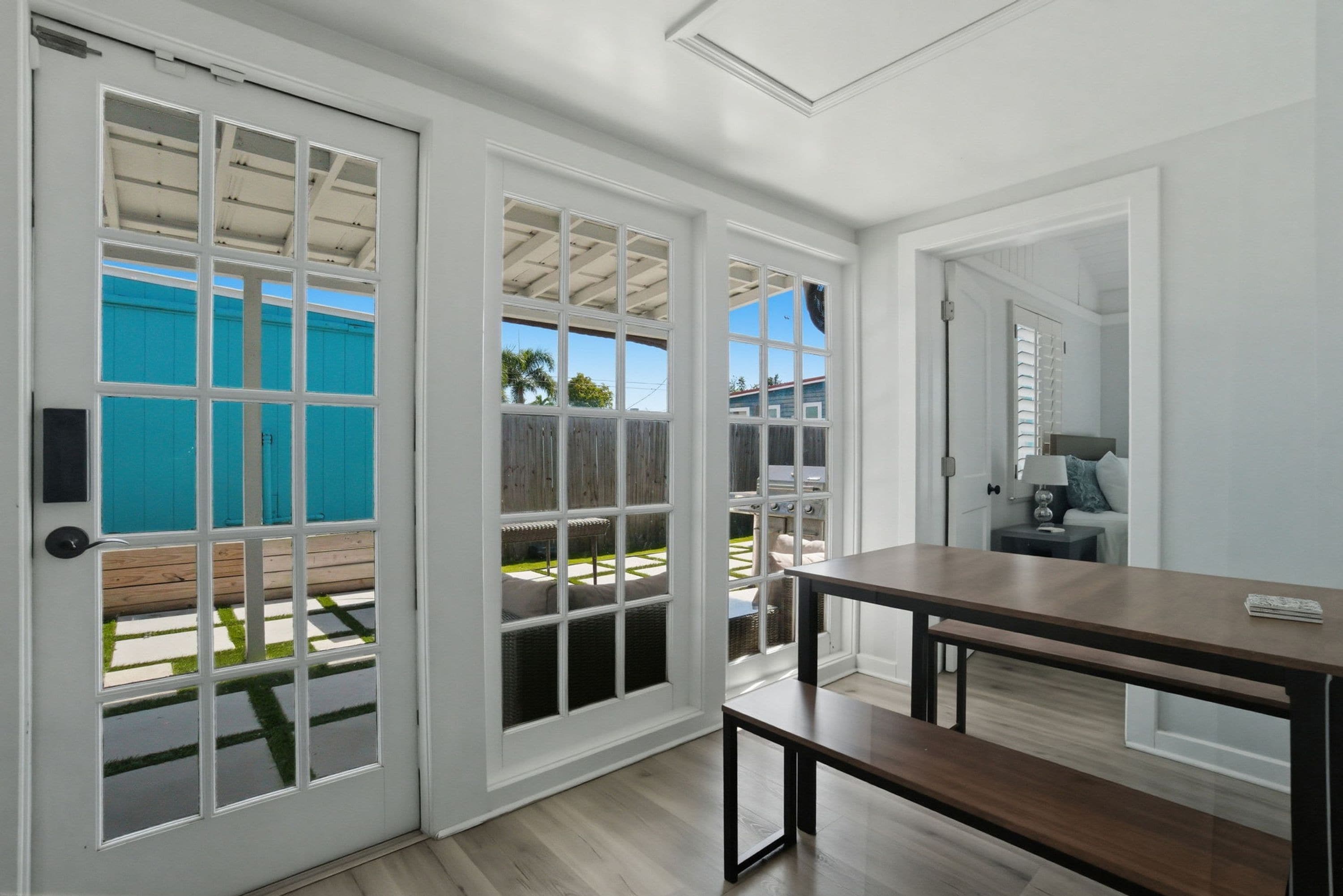 Una habitación con piso de madera, una mesa de madera oscura y un banco. Se observan ventanas con marco blanco y una puerta.