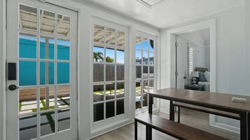 Una habitación con piso de madera, una mesa de madera oscura y un banco. Se observan ventanas con marco blanco y una puerta.