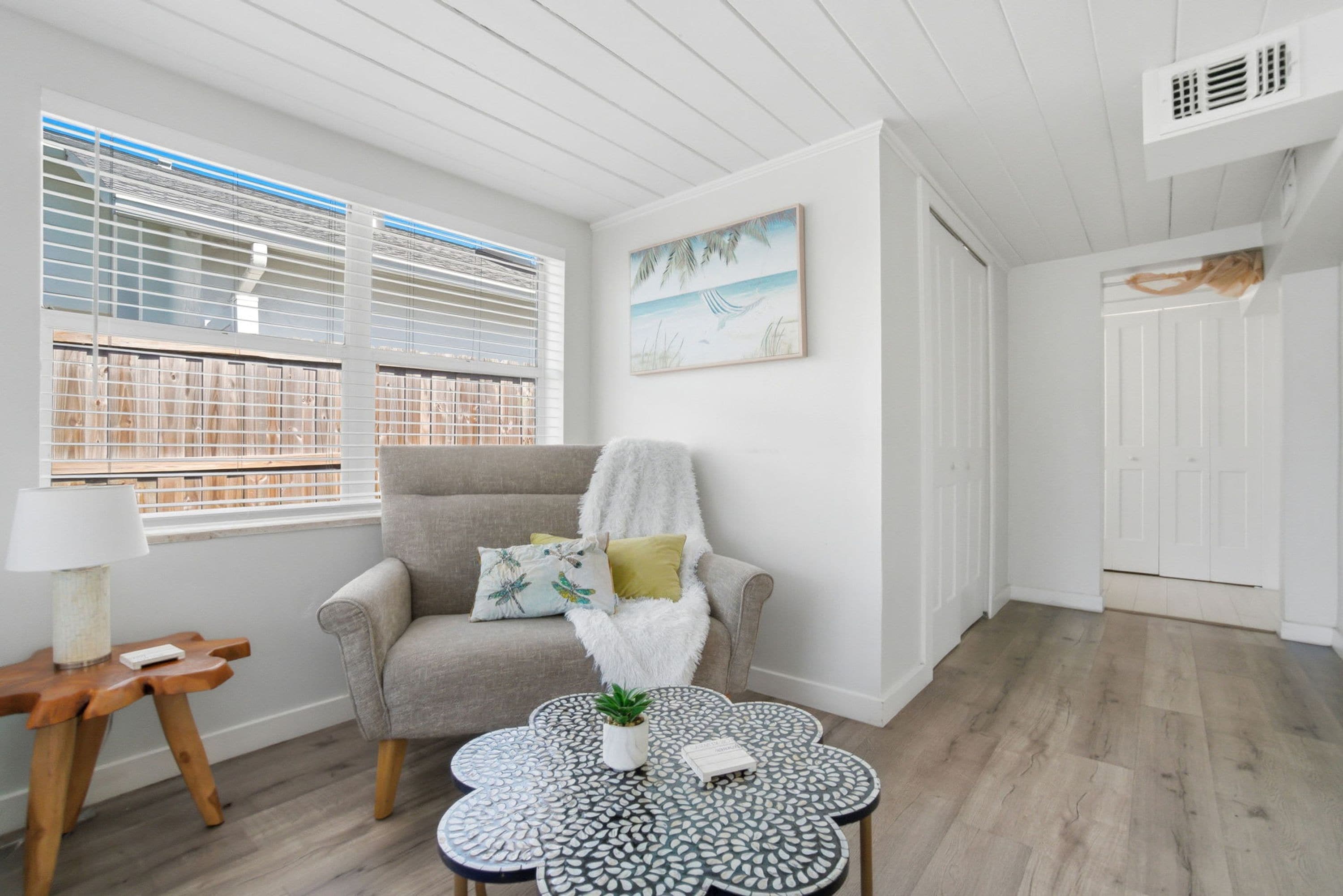 Una habitación con un sillón gris, un plaid blanco y mullido, una mesita con una planta y un cuadro de una playa.