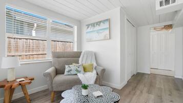 Una habitación con un sillón gris, un plaid blanco y mullido, una mesita con una planta y un cuadro de una playa.
