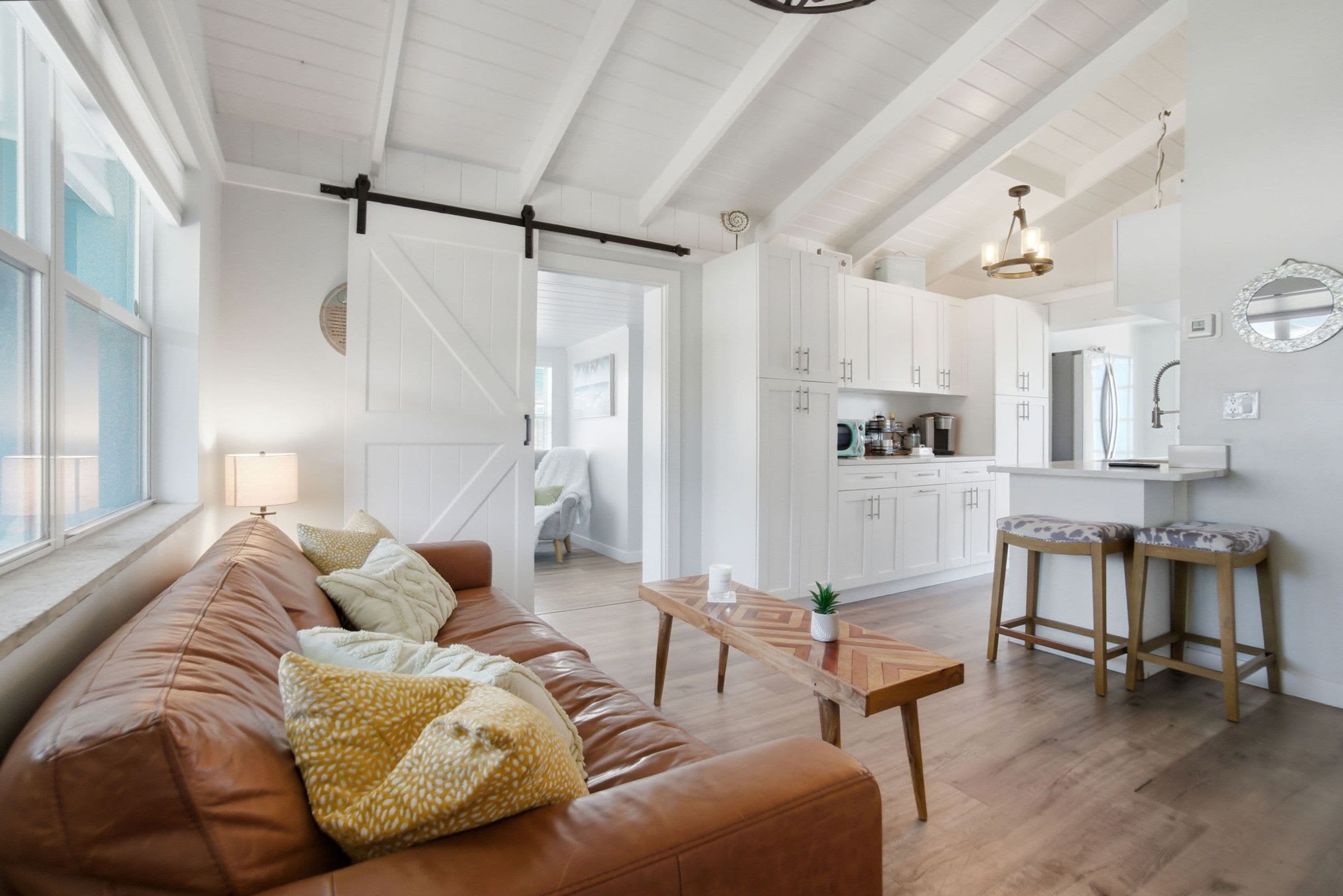 A spacious living area with a brown leather sofa, a wooden coffee table, and a kitchen with white cabinets and a bar stool.