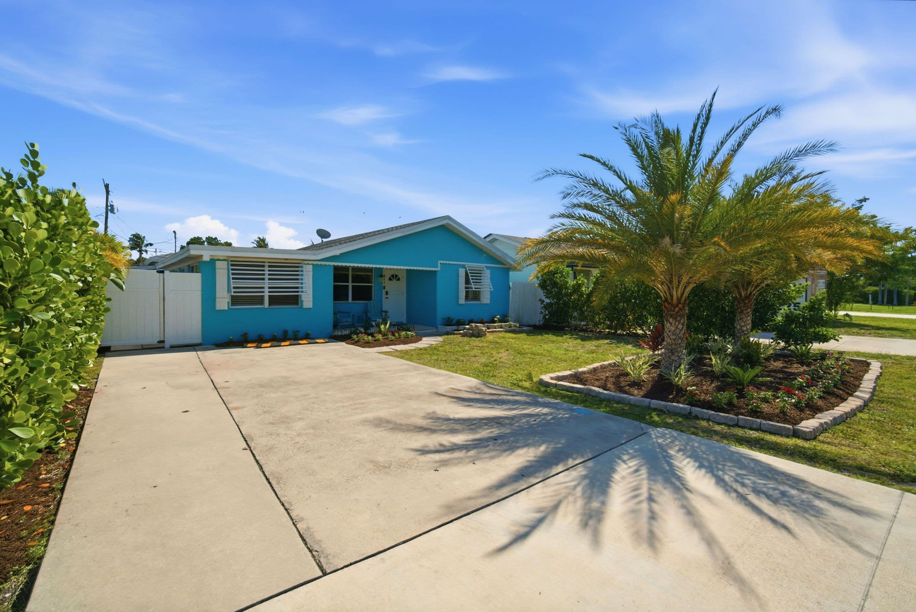 Una casa de un solo piso con fachada azul, un jardín delantero con una palmera y un cielo despejado.