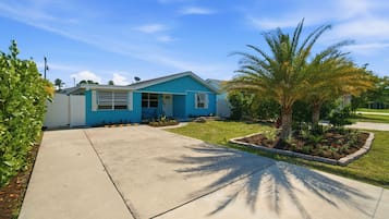 Una casa de un solo piso con fachada azul, un jardín delantero con una palmera y un cielo despejado.