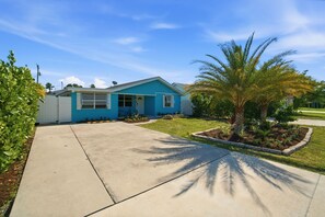 Una casa de un solo piso con fachada azul, un jardín delantero con una palmera y un cielo despejado.