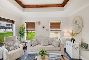 A living room with a beige sofa, a round coffee table, and a chair with star-patterned cushions. There are two windows with blinds, a wall-mounted decorative item, and a painting. - Oldsmar Escape Near Tampa Bay Safety Harbor (Oldsmar)