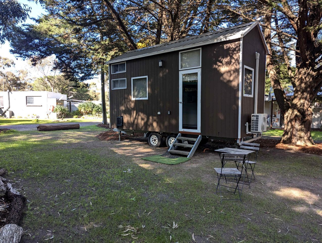 A small, single-story mobile home with a ramp leading to the door, situated in a grassy area with trees in the background.