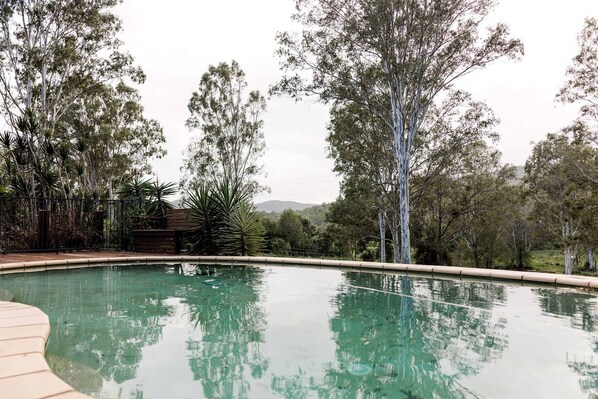 A swimming pool with a wooden deck, surrounded by trees and a hilly landscape.