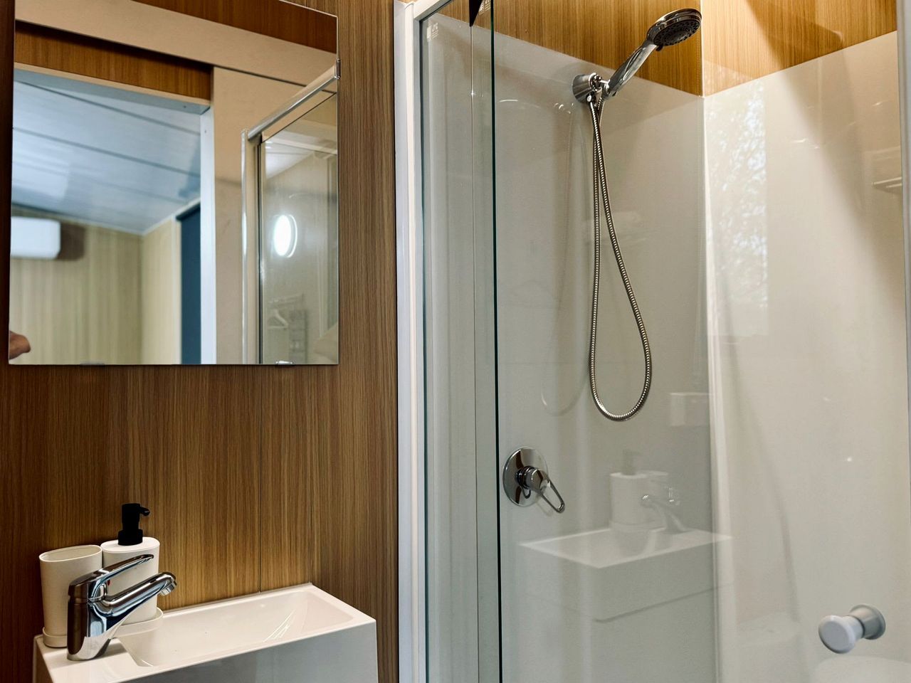 Ein modernes Badezimmer mit einer gläsernen Duschkabine, einem weißen Waschbecken mit Seifenspender und einem Spiegel darüber.