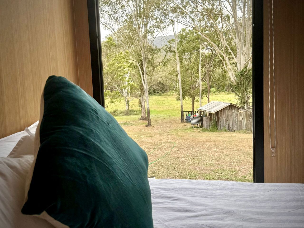 A bedroom with a bed, a pillow, and a view of a grassy area with trees and a wooden structure.