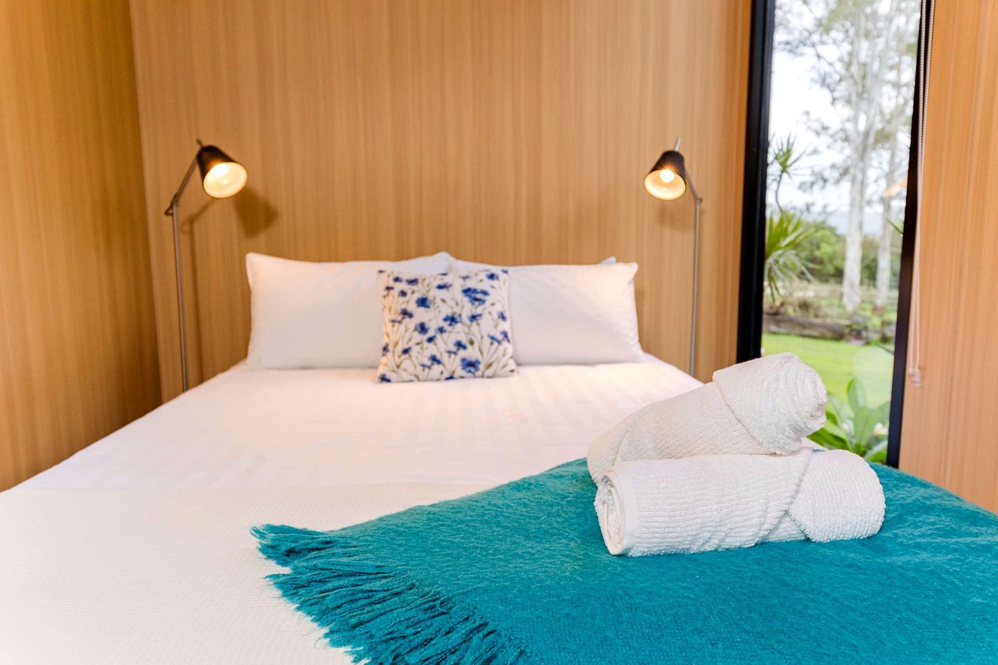 Una cama con ropa de cama blanca, una almohada floral azul y una toalla blanca enrollada sobre una alfombra color turquesa.
