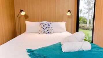 Una cama con ropa de cama blanca, una almohada floral azul y una toalla blanca enrollada sobre una alfombra color turquesa.