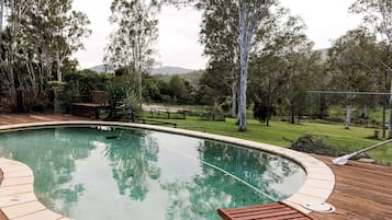 A swimming pool with a wooden deck and lounge chairs, surrounded by trees and a grassy area.