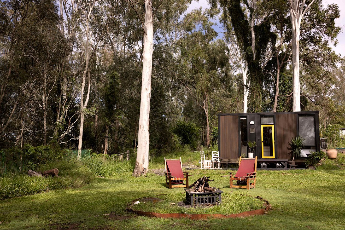 Eine moderne Holzhütte mit Tür, umgeben von einer Rasenfläche mit zwei Stühlen und einer Feuerstelle.