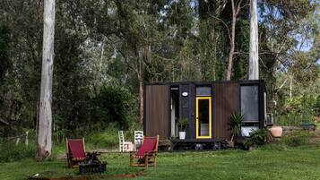 Une petite cabane avec une porte, deux chaises et une table, située dans un espace verdoyant bordé d’arbres.