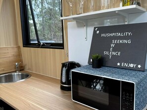 A kitchen with a microwave, a kettle, and a sink. There's a sign with the words 'HUSHPITALITY: SEEKING WISE SILENCE ENJOY'.