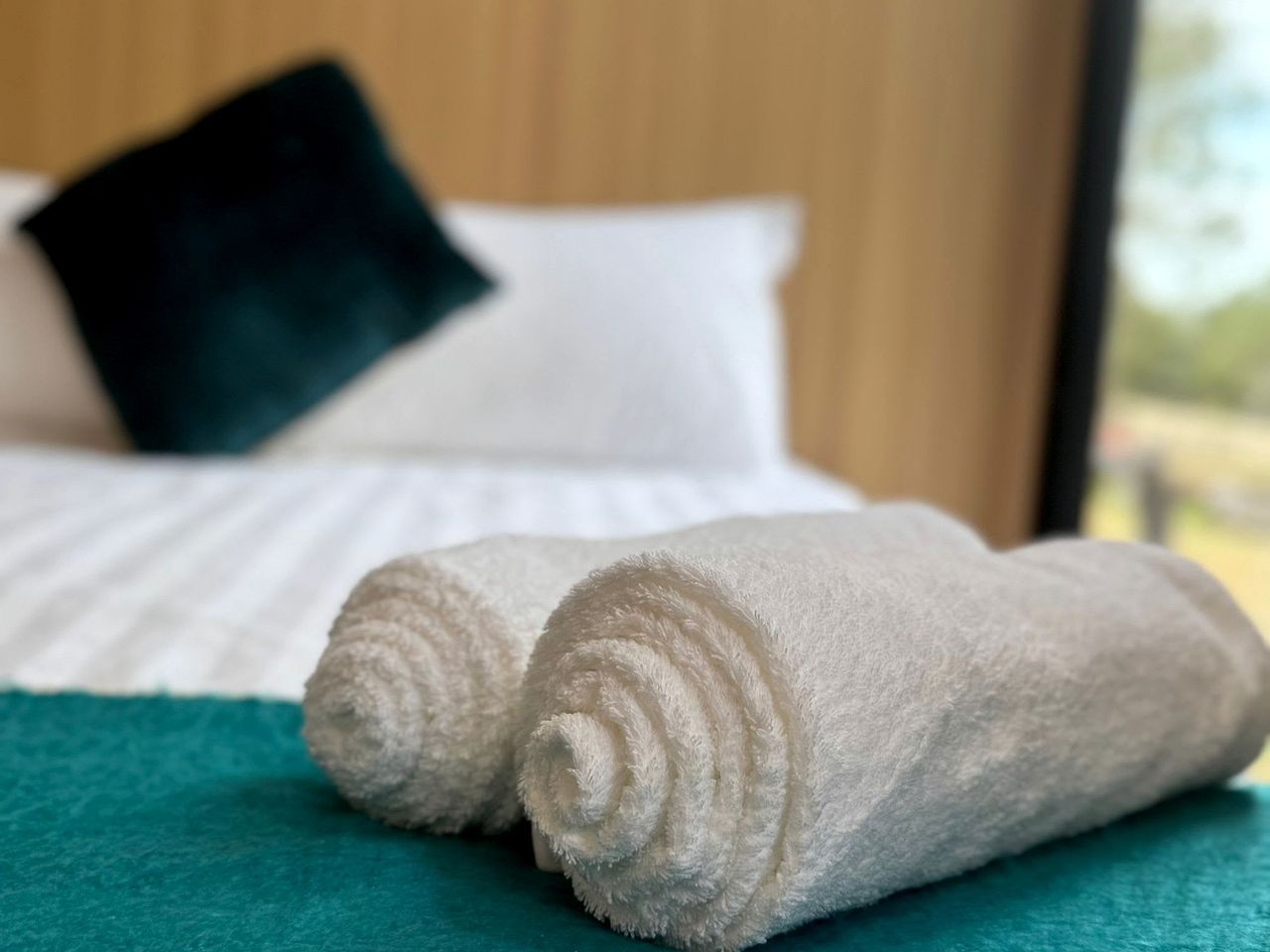 Cama bem arrumada com lençóis brancos e um cobertor azul-esverdeado, um travesseiro preto e uma janela com vista para o verde.