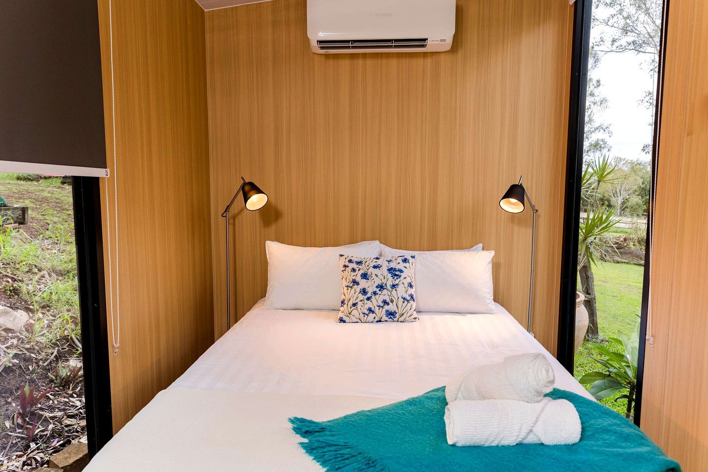 Un lit simple avec une literie blanche et un coussin à motifs floraux bleus, flanqué de deux lampes fixées au mur, dans une pièce aux murs lambrissés et offrant une vue sur la verdure à l’extérieur.