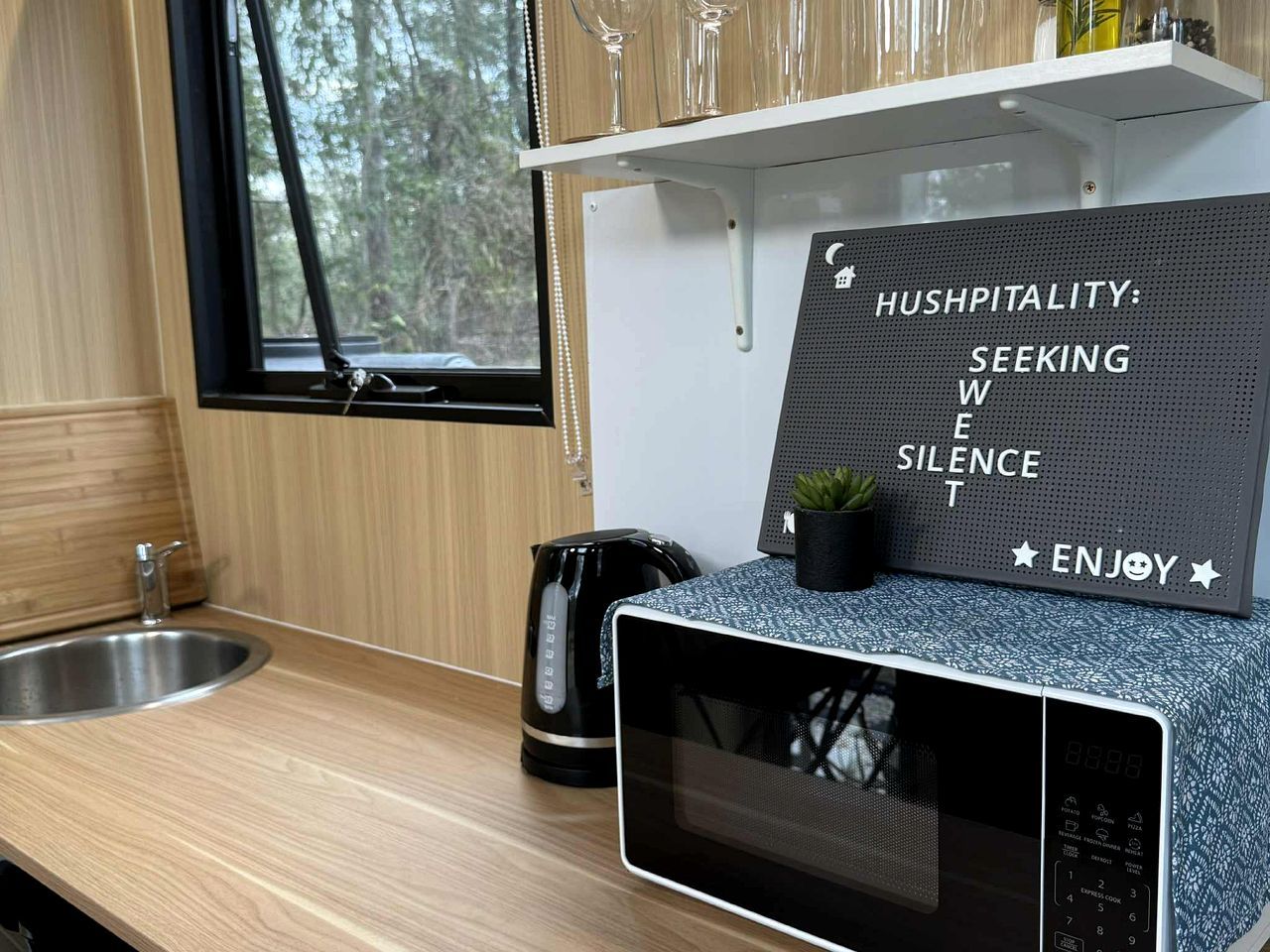 A kitchen with a microwave, a kettle, and a sink. There's a sign with the words 'HUSHPITALITY: SEEKING WISE SILENCE ENJOY'.